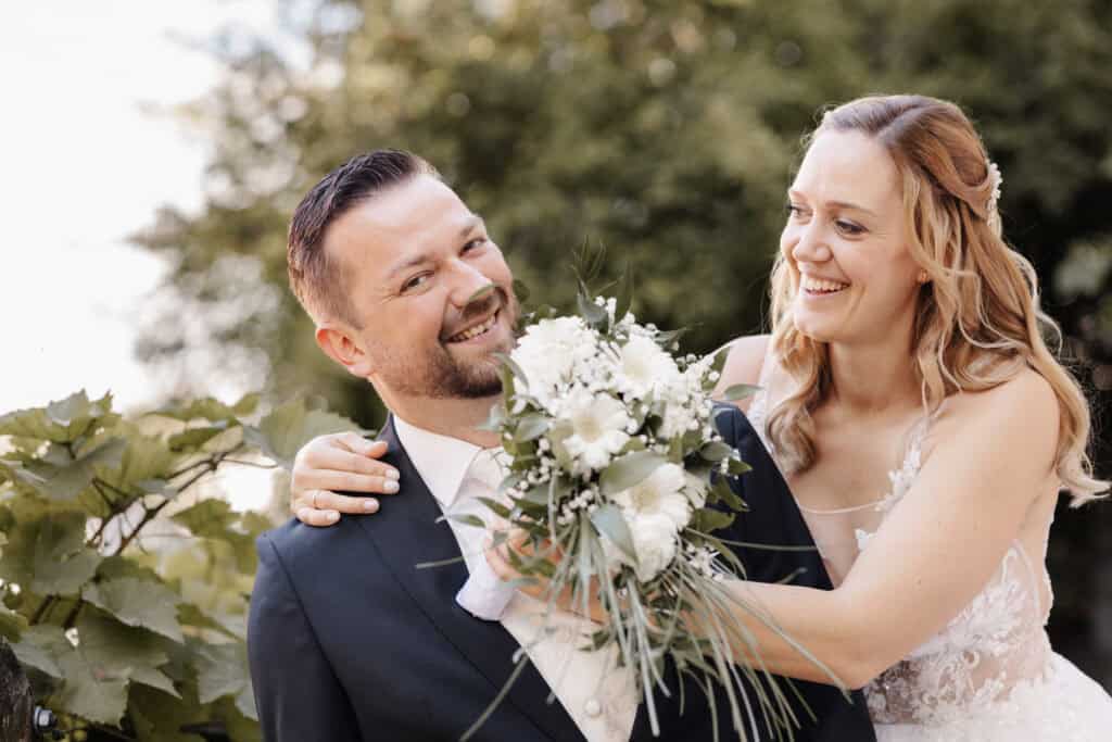 Braut und Bräutigam lachen bei einem Paarfoto im Freien, während die Braut dem Bräutigam den Brautstrauß vors Gesicht hält, festgehalten als natürlicher Moment der Hochzeitsreportage